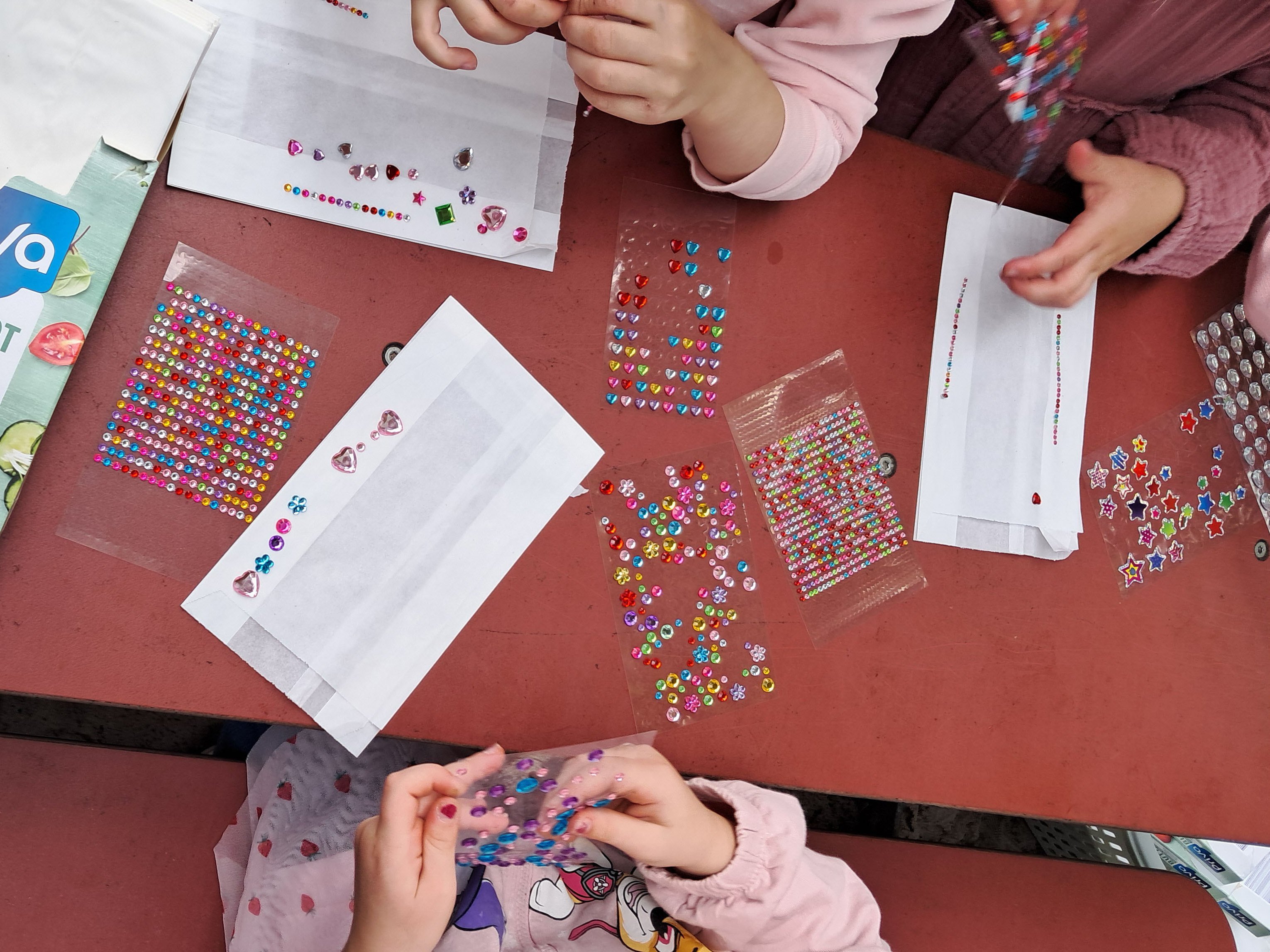 Mehrere Kinder verzieren weiße Papiertüten mit bunten Schmucksteinen.